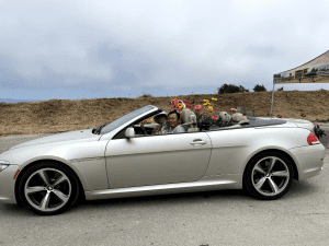 Silver convertible with a driver and passenger with a back seat filled with daisies