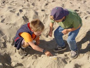 Two kids play in the sand, pointing at something buried within