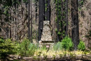 A lone stone chimney sits, fenced off among burnt redwood trees.