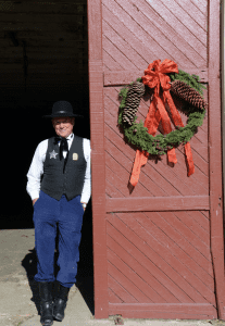 A man dressed in a black vest and old-timey hat leans against a barn door with a wreath on it