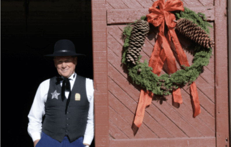 A man dressed in a black vest and old-timey hat leans against a barn door with a wreath on it